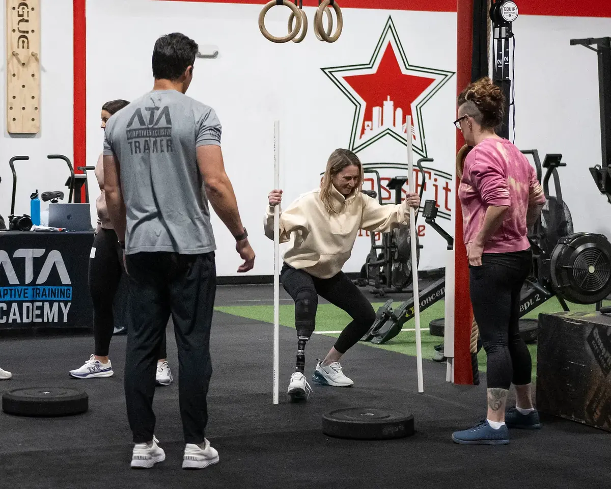 An ATA trainer coaches an athlete with a leg prosthesis through a squat using a training dowel