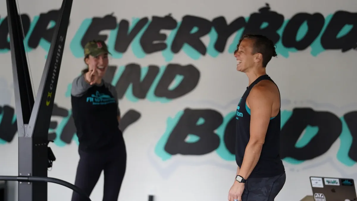 Two trainers laughing in front of a gym wall painted with the words for everybody and every body