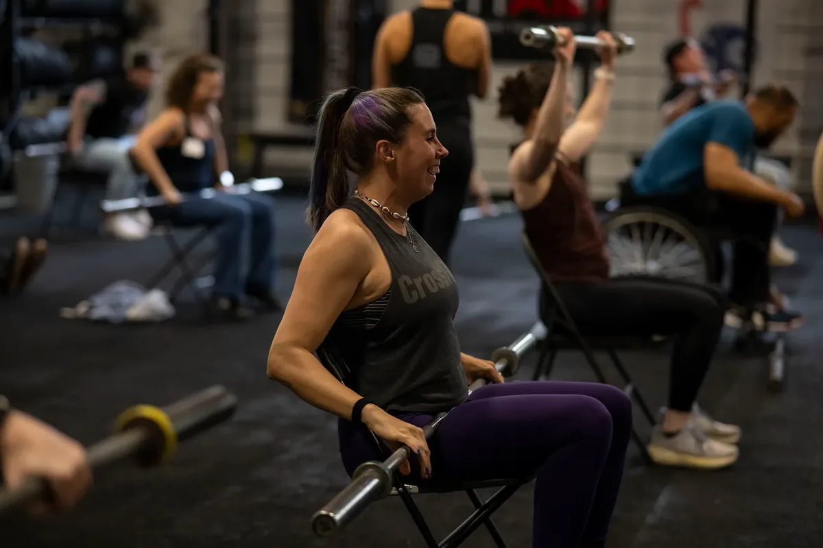 A seated athlete presses a barbell overhead during an adaptive fitness class