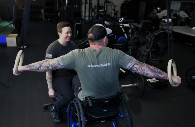An athlete trains on gymnastic rings from his wheelchair while a trainer spots him